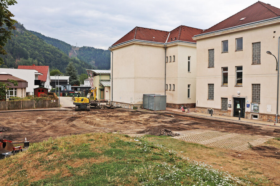 Bagger auf Baustelle mit Gebäuden und bewaldetem Hang im Hintergrund